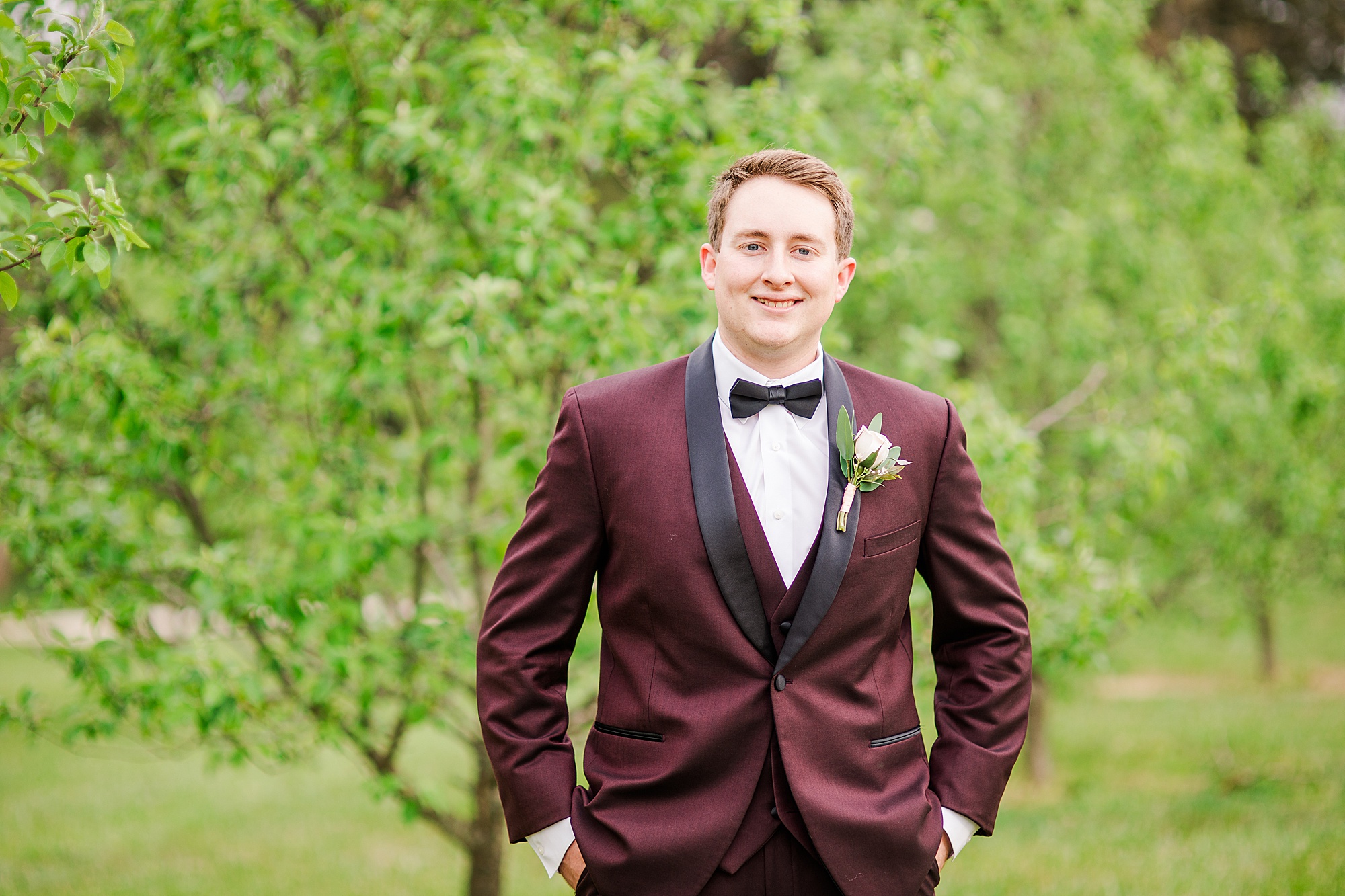 groom poses in burgundy suit in orchard at Pierce Century Farm