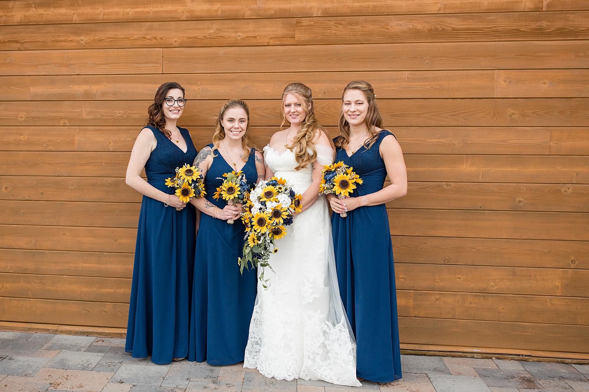 bride and bridesmaids in navy gowns with sunflowers pose outside Quail Ridge Lodge