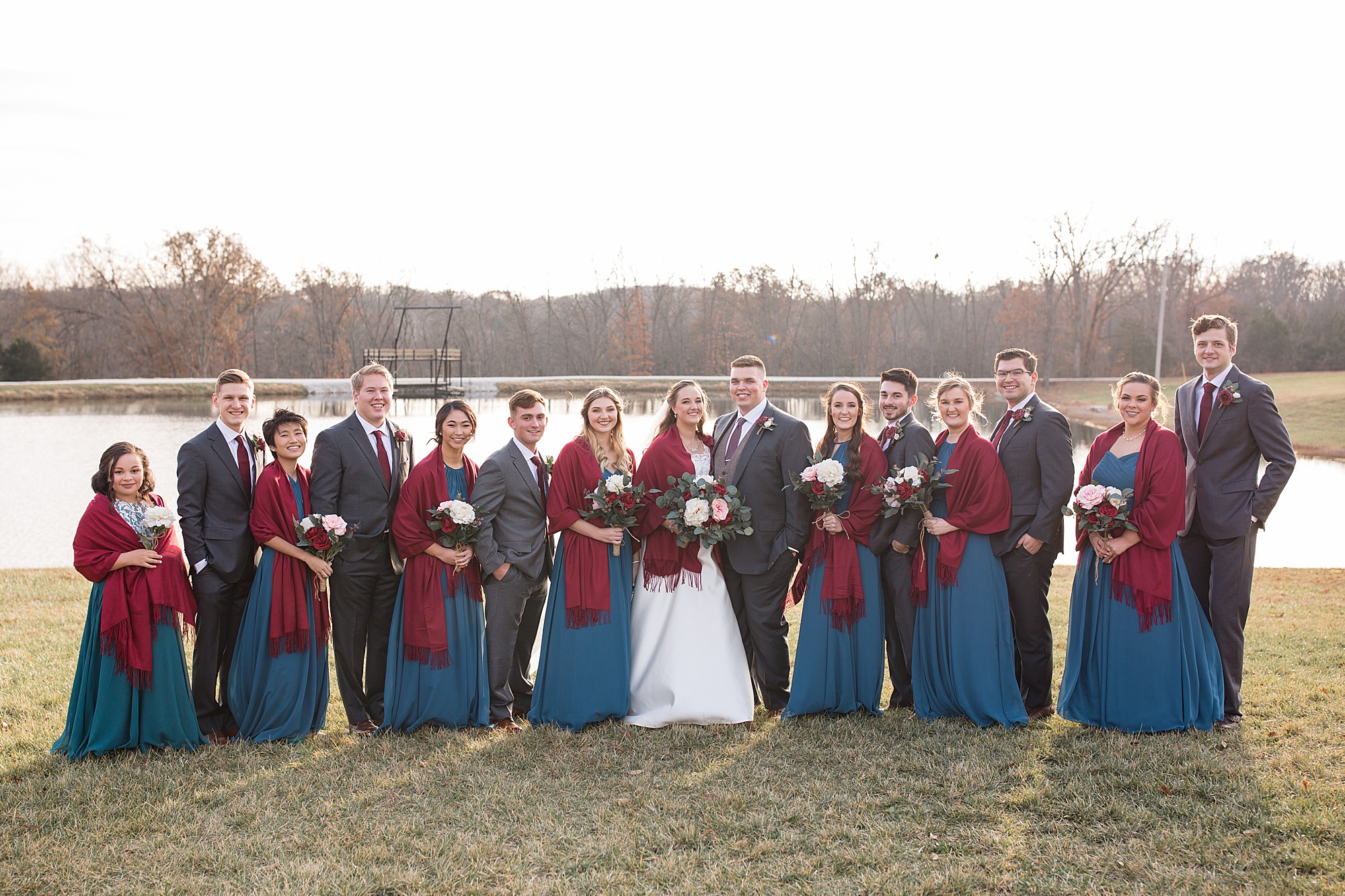 bride and groom pose with wedding party in teal gowns with red wraps and grey suits