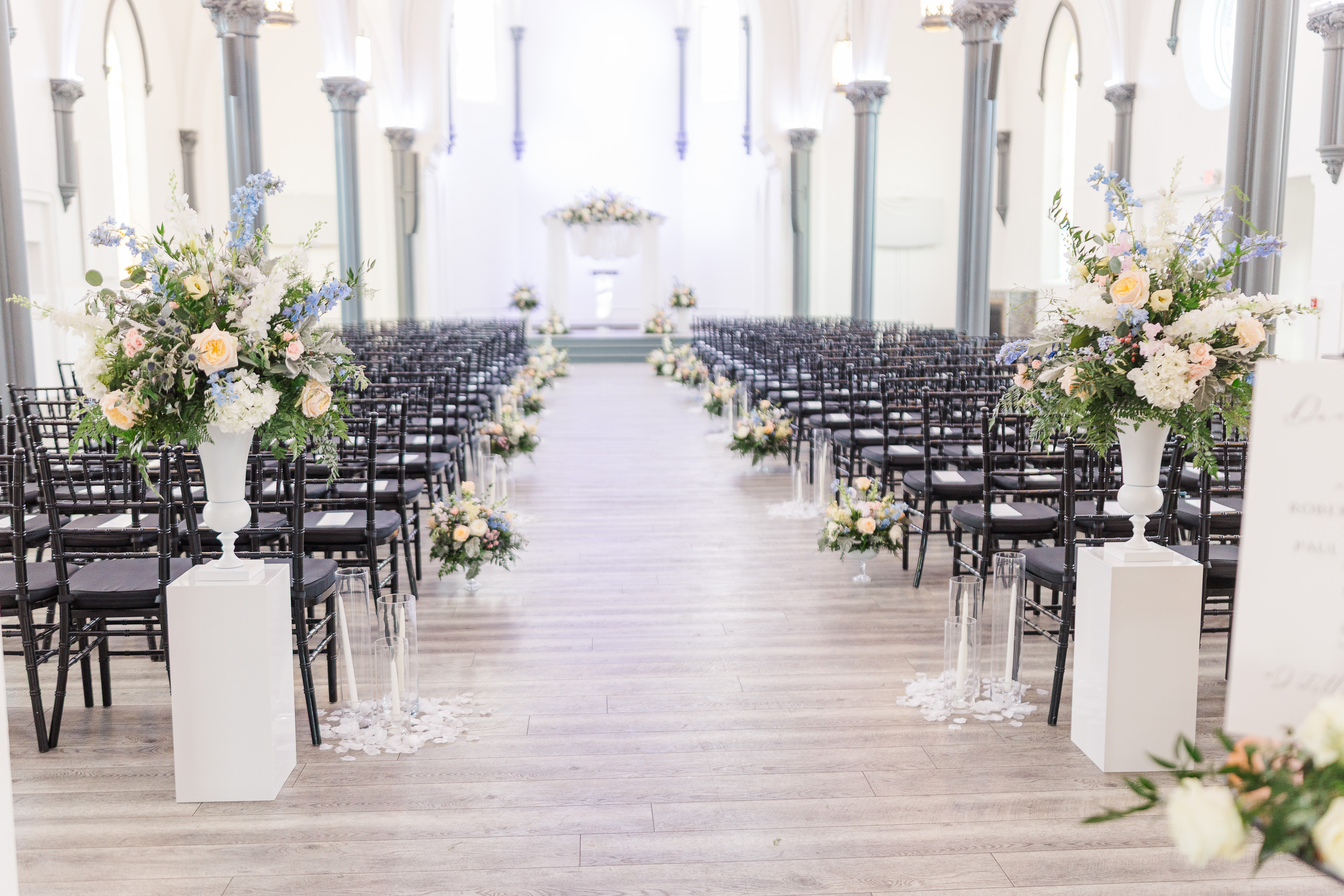 Elegant wedding ceremony setup at Main Street Abbey with black chairs, tall floral arrangements, and soft candlelight lining the aisle in a white historic chapel