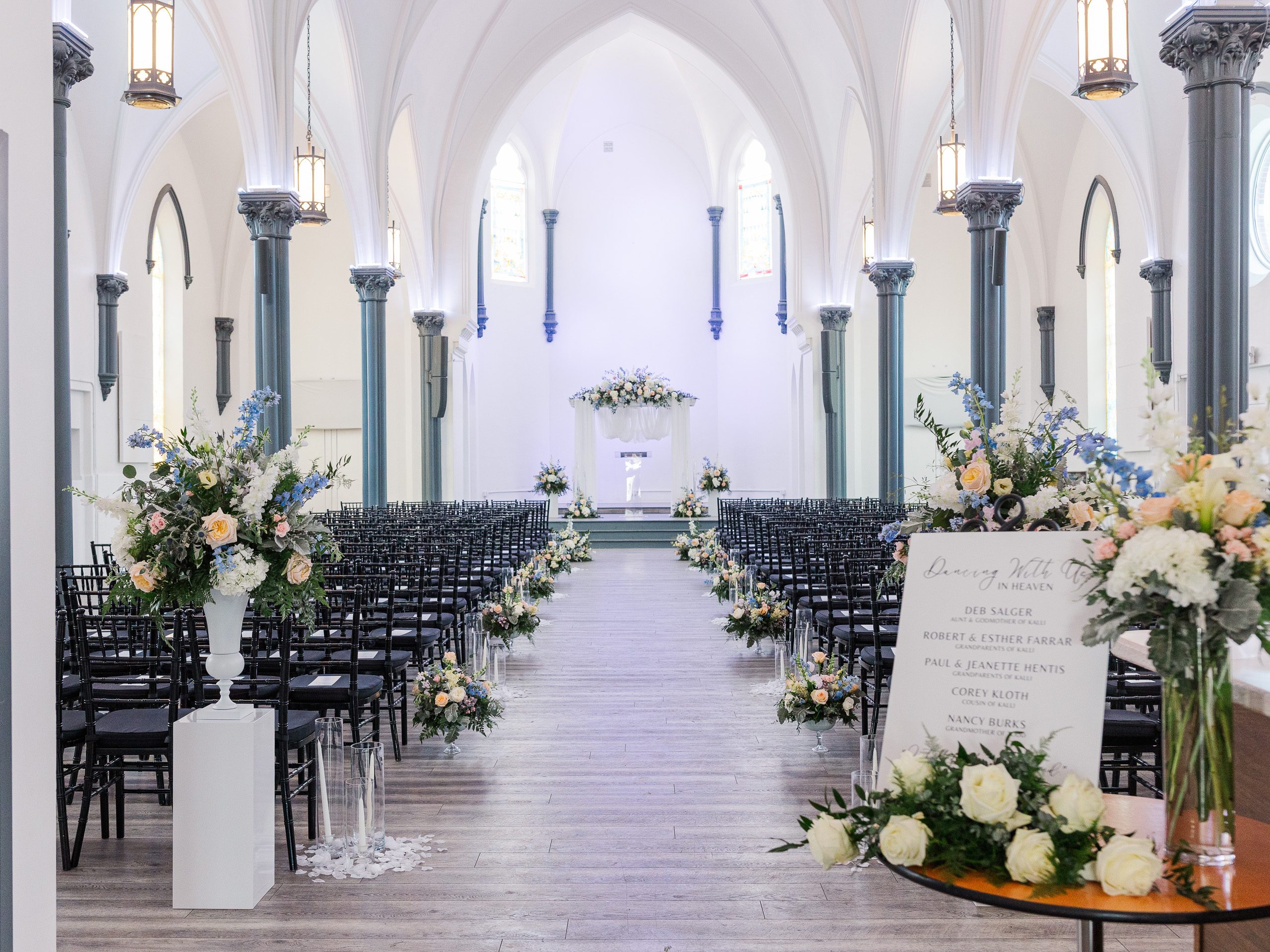 Elegant wedding ceremony setup at Main Street Abbey with black chairs, tall floral arrangements, and soft candlelight lining the aisle in a white historic chapel.