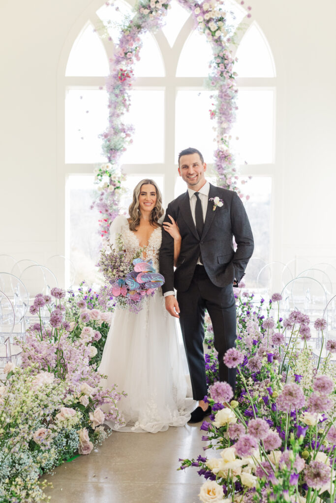Bride and groom standing beneath a floral arch of pastel purple and blush blooms inside Westwind Hills surrounded by a dramatic aisle of lush spring flowers.