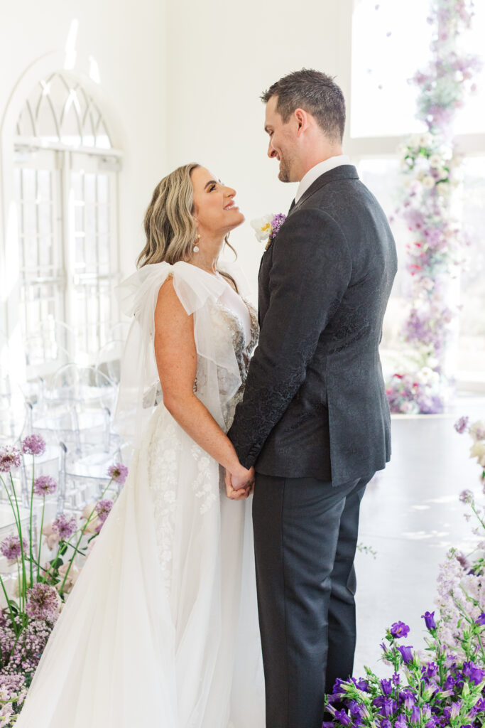 A joyful bride and groom holding hands and smiling at each other during their indoor ceremony at Westwind Hills, surrounded by lush purple florals and soft natural light.