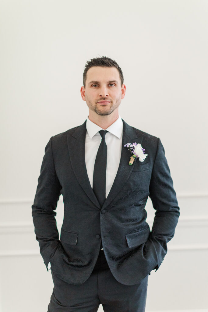 Close-up of the groom’s black textured suit jacket with a modern white and purple boutonniere, captured at Westwind Hills in Pacific, Missouri.