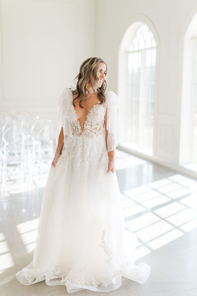 Elegant bridal portrait featuring a bride in a flowing lace and tulle gown, standing in the sunlit ceremony space at Westwind Hills with arched windows in the background.